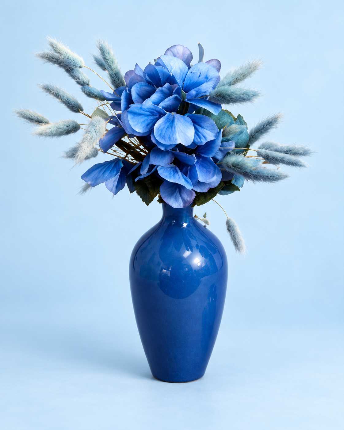 Blue flowers and soft blue grasses in a cobalt vase on a light pastel blue background