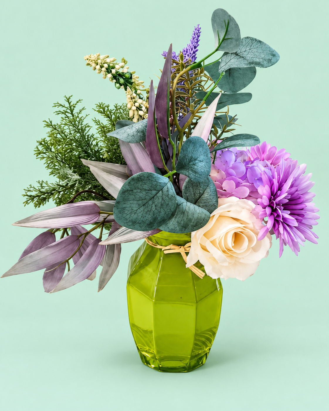 Cream and purple artificial flowers with eucalyptus in an upright green glass vase on a plain mint surface