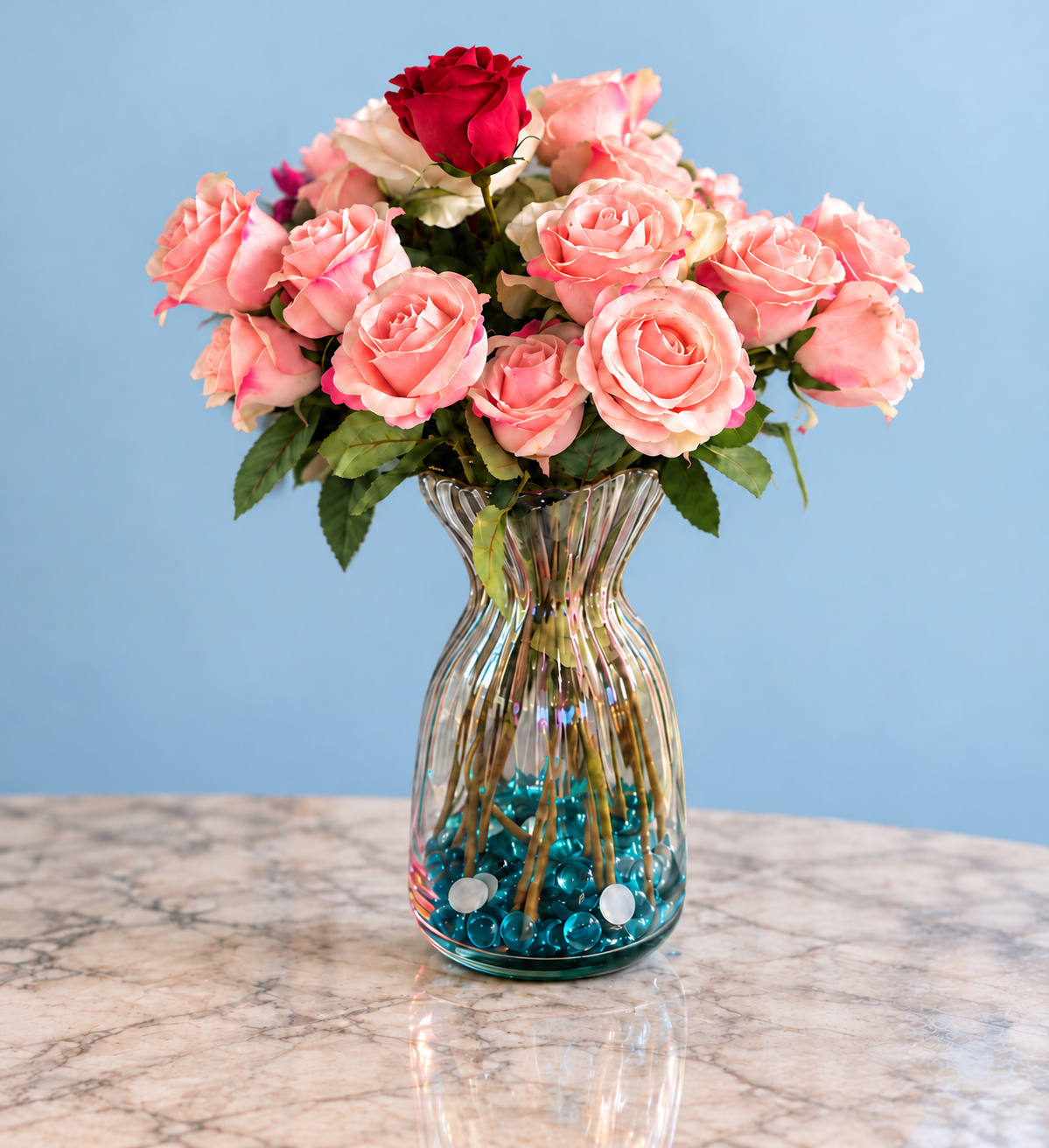 Pink roses and one red rose in a clear fluted vase with blue pebbles, on a marble table against a plain powder-blue background