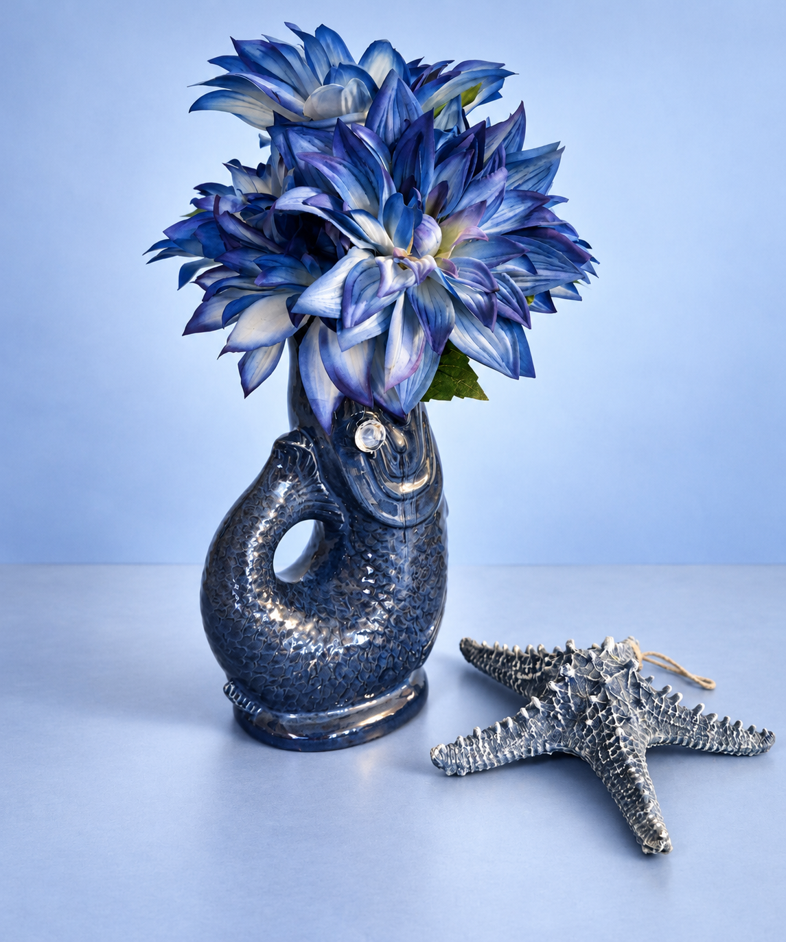 Blue artificial flowers in a glossy dark blue fish-shaped vase beside a blue-gray starfish against a powder-blue background